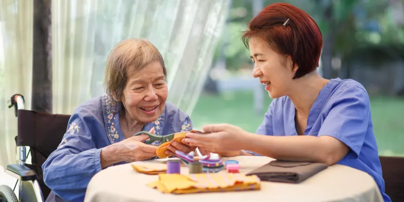 older woman using scissors with the support of an aide wearing scrubs