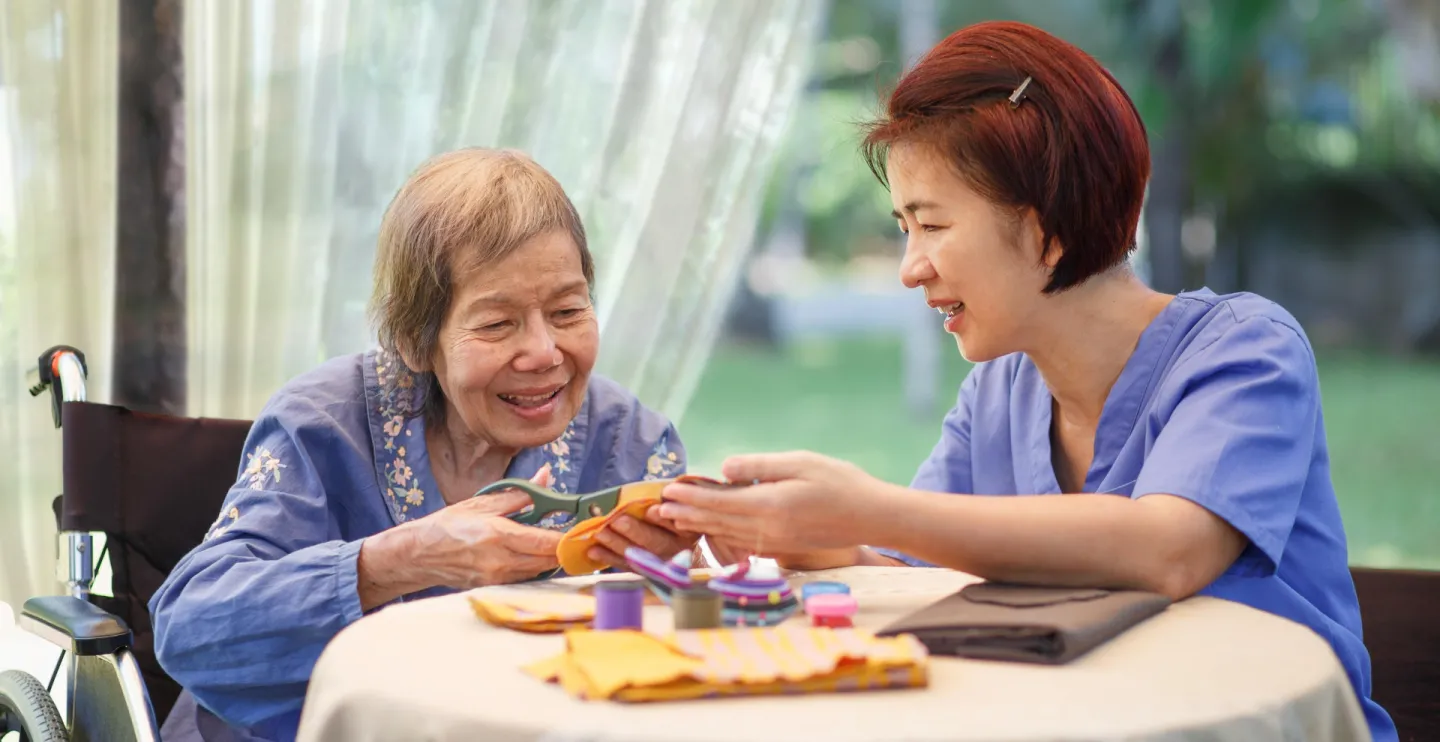 older woman using scissors with the support of an aide wearing scrubs