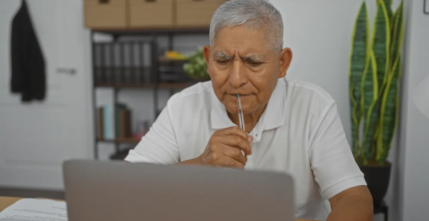 A middle-aged hispanic man in an office setting, thoughtfully looking at his laptop while holding a pen to his mouth