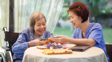 Older Asian woman at left with Asian woman caregiver who is helping her make crafts as they both sit at a table.