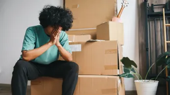 Man with dark curly hair sitting with his face in his hands in front of a pile of cardboard boxes as if stressed out about moving.